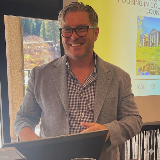 A smiling man wearing glasses, a gray blazer, and a checkered shirt stands in front of a presentation screen. The screen shows a slide with text and an image of houses in a scenic area.